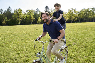 Lächelnder Vater und Sohn genießen eine Fahrradtour im Gras an einem sonnigen Tag - AUF00521