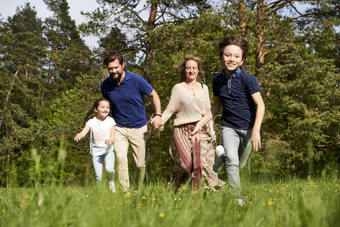 Lächelnde Familie läuft auf Gras gegen Bäume an einem sonnigen Tag - AUF00526