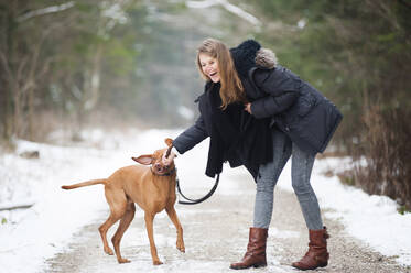 Fröhliche junge Frau spielt mit Hund auf Straße in Wald im Winter - DIGF12651