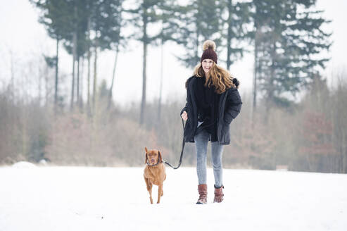 Lächelnde junge Frau, die mit ihrem Hund im Schnee vor Bäumen im Wald spazieren geht - DIGF12654