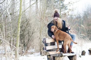 Fröhliche junge Frau mit Hund auf einer Bank sitzend vor kahlen Bäumen im Wald im Winter - DIGF12655