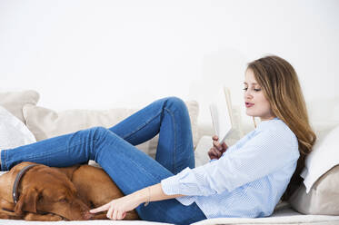 Verspielte junge Frau, die die Schnauze ihres Hundes berührt, während sie mit einem Buch auf dem Sofa zu Hause sitzt - DIGF12656