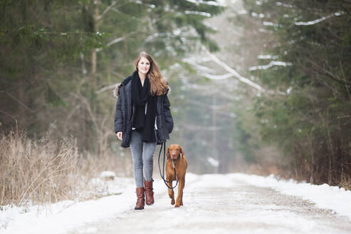 Schöne junge Frau geht mit Hund auf der Straße inmitten von Bäumen im Wald im Winter - DIGF12659