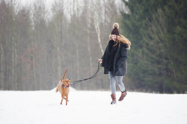 Glückliche junge Frau läuft mit Hund auf Schnee gegen Bäume im Wald im Winter - DIGF12661