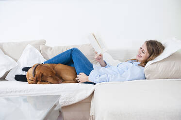 Lächelnde junge Frau liegt mit Buch und Hund auf dem Sofa im Wohnzimmer zu Hause - DIGF12662