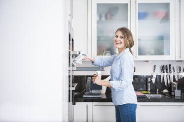 Happy beautiful young woman with long brown hair removing tray from oven - DIGF12665