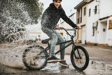 Selbstbewusster Jugendlicher, der beim Radfahren auf der Straße in der Regenzeit Wasser in eine Pfütze spritzt - ACPF00722