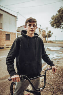 Confident teenage boy standing with bicycle on street during rainy season - ACPF00733