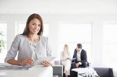 Happy businesswoman holding digital tablet while colleagues discussing in background at office - DIGF12674