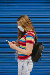 Young woman with protective mask and backpack looking at cell phone in front of blue background - AGGF00066