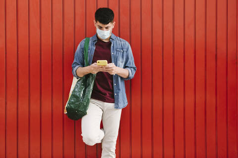 Mann mit Schutzmaske und Einkaufstasche schaut auf Handy vor rotem Hintergrund, lizenzfreies Stockfoto