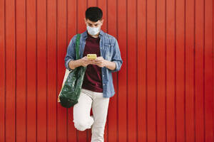 Man with protective mask and shopping bag looking at cell phone in front of red background - AGGF00069
