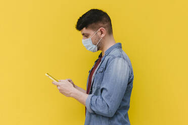 Man with protective mask looking at cell phone in front of yellow background - AGGF00071