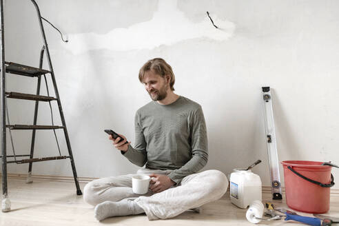 Glücklicher Mann, der sein Smartphone benutzt, während er eine Pause vom Malen zu Hause macht - EYAF01086