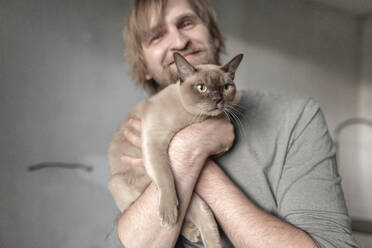 Happy man carrying Burmese cat during home renovation - EYAF01088