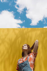 Relaxed young woman with hand in hair standing over yellow wall against cloudy sky - AFVF06438