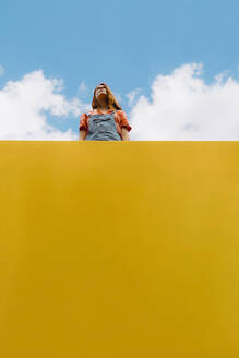 Young woman standing above yellow wall against cloudy sky - AFVF06439
