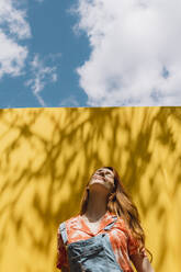 Relaxed young woman standing over yellow wall against cloudy sky - AFVF06440