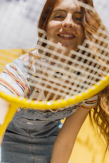Glückliche junge Frau, die an einem sonnigen Tag durch einen Badmintonschläger schaut - AFVF06442