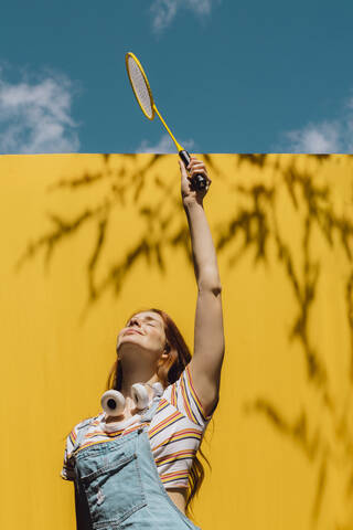 Junge Frau hält Badmintonschläger über gelbe Wand während sonnigen Tag, lizenzfreies Stockfoto