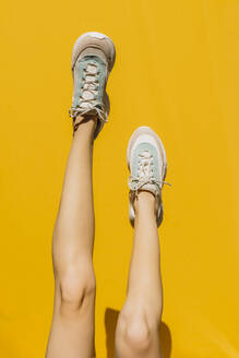 Woman's legs in sports shoes over yellow wall during sunny day - AFVF06457