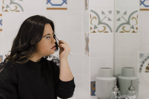 Junge Frau mit Down-Syndrom beim Schminken vor dem Badezimmerspiegel zu Hause, lizenzfreies Stockfoto