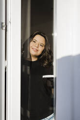 Nachdenkliche Frau mit Down-Syndrom lächelt, während sie durch das Fenster ihres Hauses schaut - DCRF00217