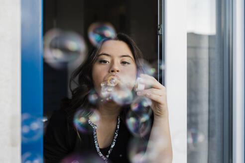 Young woman with down syndrome blowing bubbles from window at home - DCRF00231