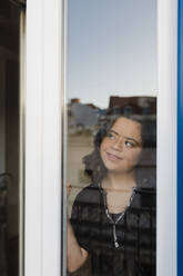 Nachdenkliche Frau mit Down-Syndrom lächelt beim Blick durch das Fenster - DCRF00232