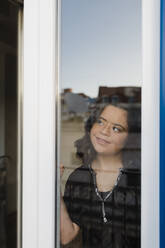 Thoughtful woman with down syndrome smiling while looking through window - DCRF00232