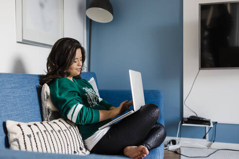 Junge Frau mit Down-Syndrom benutzt einen Laptop, während sie zu Hause auf dem Sofa im Wohnzimmer sitzt - DCRF00235
