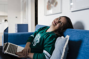 Happy young woman with down syndrome using laptop while relaxing on sofa at home - DCRF00236