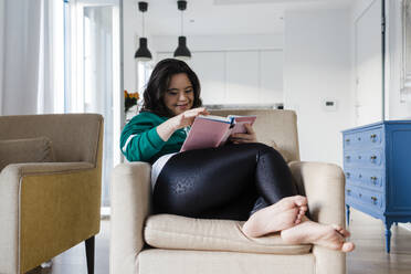 Glückliche junge Frau mit Down-Syndrom liest ein Buch, während sie zu Hause auf einem Sessel sitzt - DCRF00237