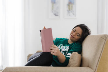 Happy young woman with down syndrome reading book while resting on armchair at home - DCRF00239