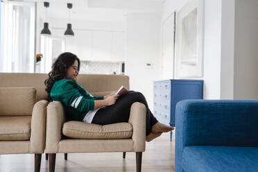 Junge Frau mit Down-Syndrom liest ein Buch, während sie zu Hause auf einem Sessel sitzt - DCRF00241