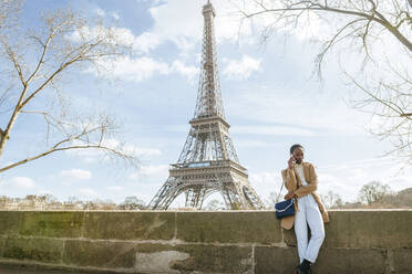 Frau, die ein Smartphone benutzt, während sie auf einer Brücke mit dem Eiffelturm im Hintergrund steht, Paris, Frankreich - KIJF03059