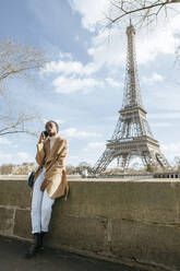 Frau, die ihr Smartphone benutzt, während sie auf einer Brücke mit dem Eiffelturm im Hintergrund gegen den Himmel steht, Paris, Frankreich - KIJF03060