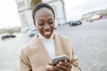 Glückliche junge Frau mit Smartphone vor dem Arc de Triomphe an einem sonnigen Tag, Paris, Frankreich - KIJF03077