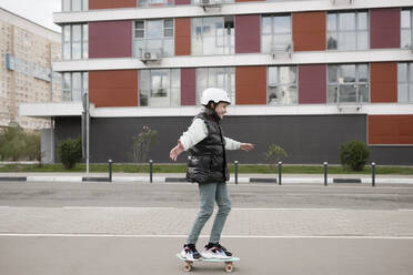 Girl wearing warm clothing skateboarding on road against building - EYAF01093