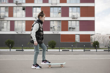 Mädchen mit Skateboard steht auf der Straße gegen Gebäude in der Stadt - EYAF01098