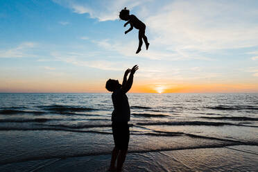 Vater wirft seine Tochter bei Sonnenuntergang im Strandurlaub in die Luft - CAVF83871