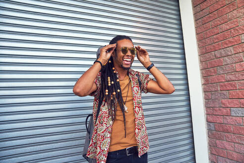 Glücklicher Mann mit Dreadlocks und Sonnenbrille vor der Garage - CAIF27844