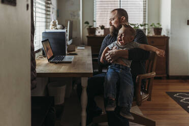 Dad working from home with one year old boy crying in his lap - CAVF84186