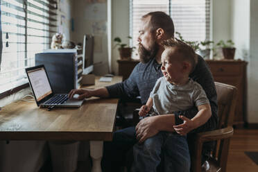 Dad working from home with one year old boy crying in his lap - CAVF84187