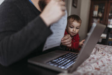 Close up of young son looking at dads computer while he works at home - CAVF84195