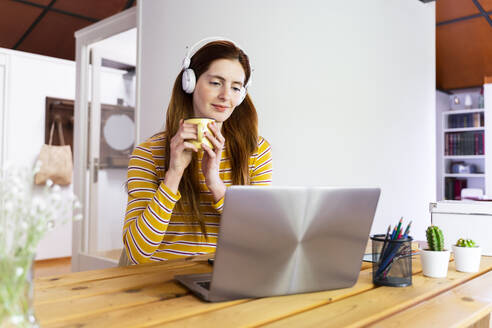 Junge Frau mit Kopfhörern, die eine Tasse hält, während sie einen Laptop auf dem Schreibtisch zu Hause benutzt - AFVF06466