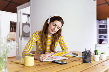 Frau mit Kopfhörern, die in ein Buch schreibt und dabei auf ihr Smartphone auf dem Schreibtisch schaut - AFVF06467