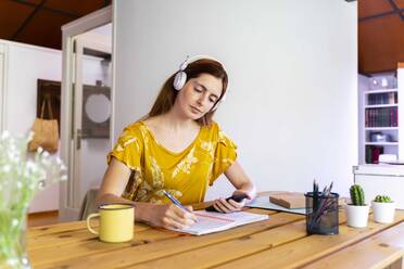 Frau hält Smartphone, während sie in einem Buch auf dem Schreibtisch zu Hause schreibt - AFVF06469