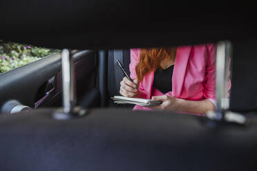 Frau schreibt in Buch, während sie im Auto sitzt, gesehen durch den Fahrzeugsitz - LJF01541