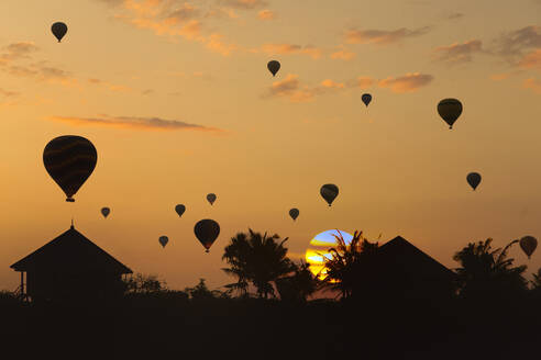 Indonesien, Bali, Silhouetten von Heißluftballons fliegen über Küstenhütten bei stimmungsvollem Sonnenuntergang - KNTF04654